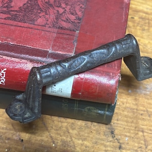 May include: A weathered, dark metal handle with a textured surface, resting on a stack of antique books with red and green covers. The handle is attached to the books with small, decorative fasteners.