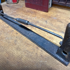 May include: An antique metal tool with a dark, aged appearance. It features a long, rectangular base with a central rod and two wooden handles. The tool appears to be used for smoothing or shaping materials.