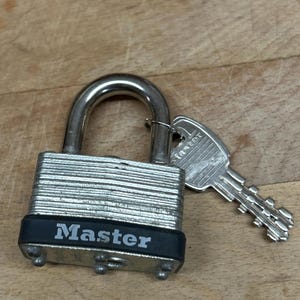 May include: A silver-colored laminated steel padlock with a U-shaped shackle and a black rubber bumper featuring the word Master in white text. A matching silver-colored metal key with the word Master embossed on the head is inserted in the shackle.
