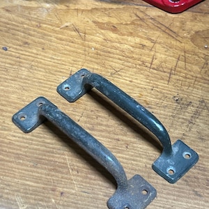 May include: Two vintage metal handles with a curved bar and square mounting plates. The handles are a dark, weathered color, showing signs of age and use. Each plate has two or three screw holes. The handles are likely for a cabinet or drawer.