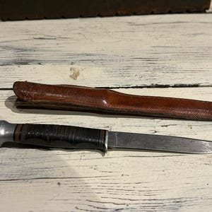 May include: A vintage hunting knife with a leather sheath. The knife has a dark handle and a silver bolster. The sheath is brown leather. The knife blade is silver and shows signs of wear.