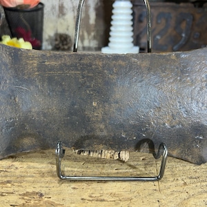 May include: A vintage, double-bit axe head with a weathered, dark brown finish. The axe head is displayed on a metal stand, showcasing its symmetrical design and aged texture. This antique tool suggests a history of use and craftsmanship.