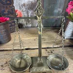 May include: A brass balance scale featuring a central vertical post with a decorative top, a square base with ornate detailing, and two circular pans suspended by metal chains. The metal has a weathered, aged patina with visible signs of oxidation.