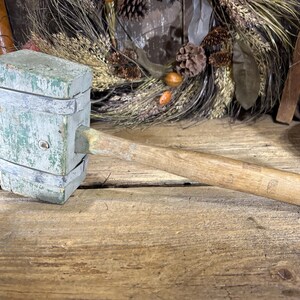 May include: An antique wooden mallet with a rectangular head painted in faded green and a long, natural wood handle. The mallet is resting on a weathered wooden surface, suggesting a vintage or rustic aesthetic.