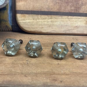 May include: Five clear glass cabinet knobs with a hexagonal shape and silver metal screws. The knobs have a slightly frosted appearance.
