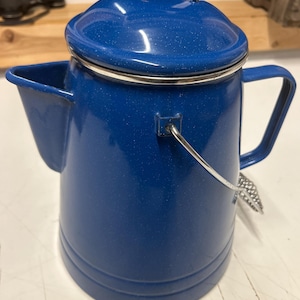 May include: A blue enamelware coffee pot with a lid and handle. The pot has a spout and a silver-colored handle. The pot is a deep blue color with a speckled finish. The pot is designed for camping or outdoor use.