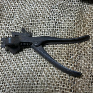 May include: A vintage, dark metal tool with a lever handle and a spring mechanism. The tool has a claw-like end and is likely used for a specific mechanical task. The object is set against a textured, neutral-toned background.
