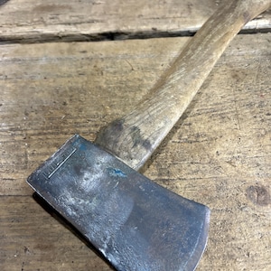 May include: A vintage hand axe with a weathered wooden handle and a dark, oxidized metal head. The metal blade features a rectangular stamp with the text "COLLINS" clearly visible on the top corner, showing signs of heavy use and patina.