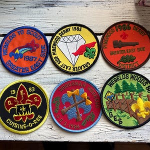 May include: Six colorful embroidered patches with various designs and text. The patches feature a diamond, a campfire, a scout symbol, a car, and trees. The text on the patches includes "Pinewood Derby", "Webelos Woods", "Greater East Side", "Cuisine-O-Ree", "Woodman Weekend", and "Scout".