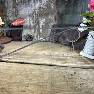 May include: A rustic, wrought iron shelf bracket with a curved design. The bracket is dark gray and appears to be handmade. It features a hook for hanging items and is mounted on a wooden surface. The bracket is designed for wall mounting.