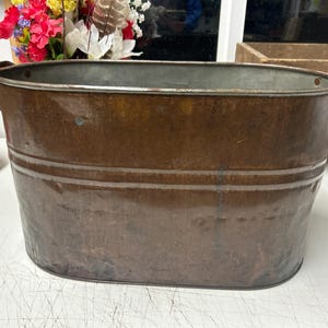 May include: A vintage, oval-shaped copper tub with two side handles. The tub has a weathered, aged appearance with visible patina and horizontal bands. The interior is a lighter color than the exterior. The tub is likely used for decorative or functional purposes.