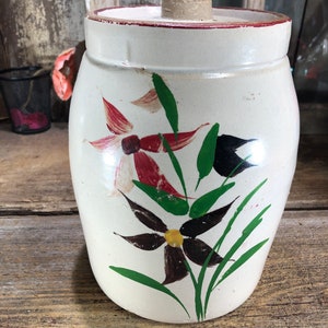 May include: White ceramic crock with a lid, hand-painted with green leaves and red, white, and black flowers.