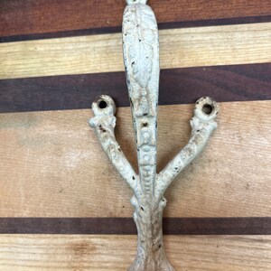 May include: White cast iron coat hook with a decorative design. The hook has three prongs and a loop handle.