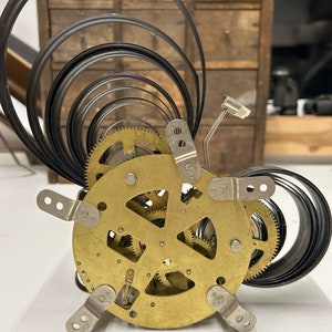 May include: Close-up of a clockwork mechanism with a brass gear system and a coiled spring. The spring is made of black metal and is coiled in a spiral shape.