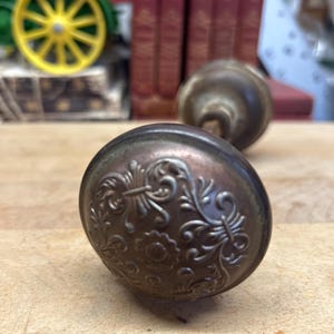May include: A round, metallic door knob with a dark, aged patina finish. The surface features an embossed, intricate floral and scrollwork pattern centered around a small flower motif. A second, matching door knob is visible in the background.