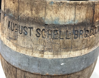 Könnte beinhalten: Ein verwittertes Holzfass mit Metallbändern. Der Text "AUGUST SCHELL BRG CO" ist in das Holz geschnitzt. Das Fass hat ein rustikales, gealtertes Aussehen mit sichtbarer Holzmaserung und einem dunklen Innenraum.