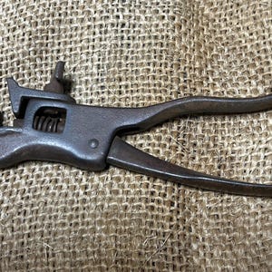 May include: A vintage, rusty metal tool with a spring mechanism and curved handles. The tool has a clamp-like head and a lever for operation. The overall design suggests it is a specialized tool, possibly for a specific craft or trade.