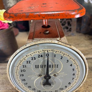 May include: Vintage Way-Rite scale with a distressed orange platform and a white and gray face. The scale features a black needle and a dial with numbers from 1 to 24. The scale is an antique kitchen tool.