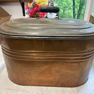 May include: A copper-colored, oval-shaped antique wash tub with a lid. The tub has a handle on the lid and decorative handles on the sides. The metal has a weathered appearance, suggesting age and use. The tub is likely used for washing clothes.