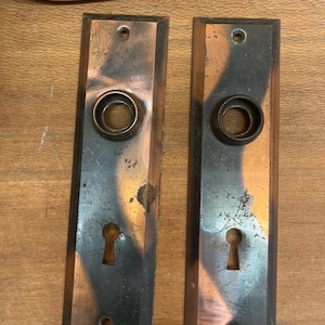 May include: Two antique door plates made of copper with a keyhole and a round knob. The plates have a distressed, aged look with a dark patina.