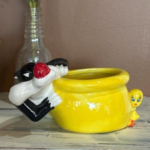 May include: A yellow ceramic planter shaped like Tweety Bird from Looney Tunes. Sylvester the Cat is peeking out from the side of the planter.