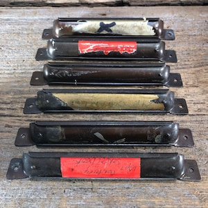 May include: Six vintage metal label holders with a dark finish are arranged on a wooden surface. Each holder has a curved design with mounting holes and a slot for a label. Some holders have remnants of labels with text and markings.