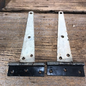 May include: Two black metal hinges with white painted metal straps. The hinges are on a wooden surface.