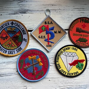 May include: A collection of vintage Boy Scout patches. The patches feature various designs, including a diamond, a campfire, axes, and a cannon. Each patch has text indicating the event and year, such as "Frontier Challenge 1985" and "Pinewood Derby 1986".