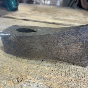 May include: A weathered, dark brown, metal axe head with a rounded edge and a hole in the center. The axe head is lying on a wooden surface.