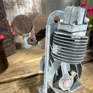 May include: A vintage metal air compressor pump featuring a silver-toned finned cylinder block, a circular pressure gauge, and a rusted brown tank. The mechanical assembly is mounted on a flat rectangular base with visible bolts and hardware.
