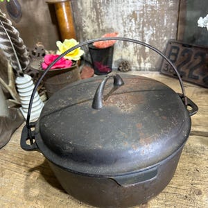 May include: A round, black cast iron Dutch oven with a matching lid and a metal bail handle. The pot features a central handle on the lid and side handles on the main body. The surface shows signs of wear, including patches of rust and discoloration.