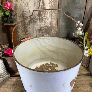 May include: A vintage white metal bucket with a rusty rim and handle. The bucket has a cylindrical shape and shows signs of wear and age, with rust spots on the exterior. The handle is made of a similar rusty metal.