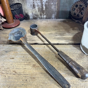 May include: Two vintage metal ladles with wooden handles. The ladles are rusty and have a worn patina. One ladle has a round bowl and the other has a long, narrow bowl.