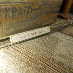 May include: A vintage metal whisk with a white label that reads "WAYNE DAIRY FEEDS". The whisk is lying on a wooden surface.