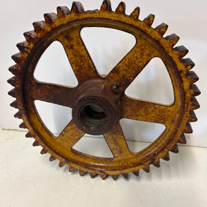 May include: A large, rusty metal gear with a yellow and brown color scheme. The gear has multiple teeth around the outer edge and several spokes connecting the center hub to the outer ring. The gear appears to be vintage or antique.
