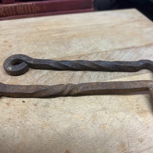 May include: Two hand-forged iron hooks with a dark, weathered, and rusted finish. Each hook features a circular eyelet at one end, a central twisted metal shaft, and a sharp, angled hook at the opposite end, showing signs of age and oxidation.