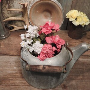 May include: A vintage aluminum teapot with a wooden handle, filled with pink, white, and red artificial flowers. The teapot is sitting on a rustic wooden surface.
