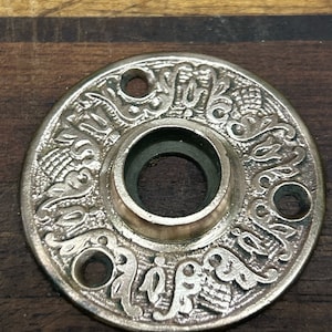 May include: A silver-toned, round door knob escutcheon with an ornate, floral design. The escutcheon has a hole in the center for the knob.