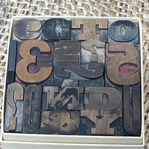 May include: A set of vintage wooden letterpress printing blocks, including numbers and symbols, arranged in a white box. The blocks are various shades of brown and black, showing signs of wear.