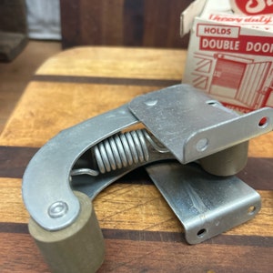 May include: Silver door stop with a spring and a rubber roller. The door stop is designed to hold a double door open.