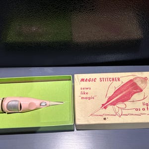 May include: A vintage Magic Stitcher sewing tool in a green box, next to its original box. The stitcher is pink and white. The box reads "MAGIC STITCHER" and "sews like 'magic' light as a feather".