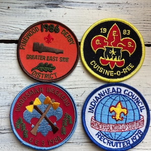 May include: Four embroidered scout patches. The top left patch is orange with a car and the text "Pinewood 1986 Derby Greater East Side District". The top right patch is black with a yellow border and the text "19 E. 83 G. S. Cuisine-O-Pee". The bottom left patch is red with a blue border and the text "Woodman Weekend CES 1984". The bottom right patch is white with a blue border and the text "Indianhead Council Wonderful World of Scouting Recruiter 1978".