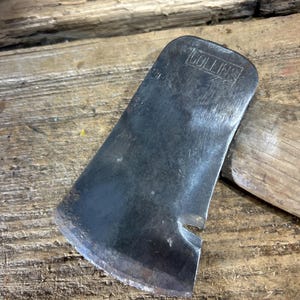 May include: A metal axe head with a curved cutting edge and a notch near the base, featuring the word COLLINS stamped in a rectangular border on the flat surface. The axe head is attached to a wooden handle, showing signs of wear and use.
