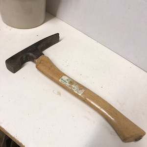May include: A vintage wooden-handled brick hammer with a metal head. The hammer has a green and white label that reads "Quality" and "Made in USA".