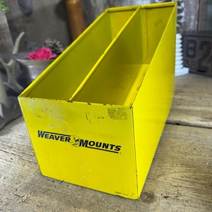 May include: A bright yellow metal organizer with two compartments. The organizer has the text "WEAVER MOUNTS" printed on the front. The item is sitting on a wooden surface.