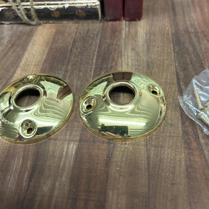 May include: Two gold-toned door knob escutcheons with a round hole in the center. The escutcheons are on a wooden surface. A small bag of screws is in the background.