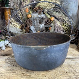 May include: A dark gray cast iron cauldron with a curved handle. The cauldron has a wide, open mouth and sits on a wooden surface. The background includes a wreath and floral arrangement.