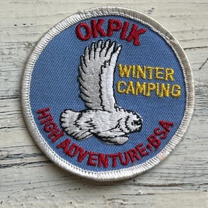 May include: A blue and white embroidered patch with the text "OKPIK WINTER CAMPING HIGH ADVENTURE BSA". The patch features a white owl with red and yellow accents.