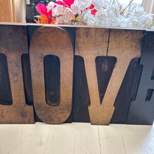 May include: A wooden block sign spelling out the word "LOVE" in large, individual letters. The letters are a mix of dark brown and lighter wood tones. The top of the sign holds an arrangement of white and pink artificial flowers.