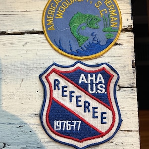May include: Two embroidered patches. The top patch is round and blue with a yellow border. It features a green fish jumping out of the water. The text on the patch reads "American Bass Fisherman Woodruff, S.C.". The bottom patch is a blue shield with a red and white diagonal stripe. The text on the patch reads "AHA U.S. Referee 1976-77".